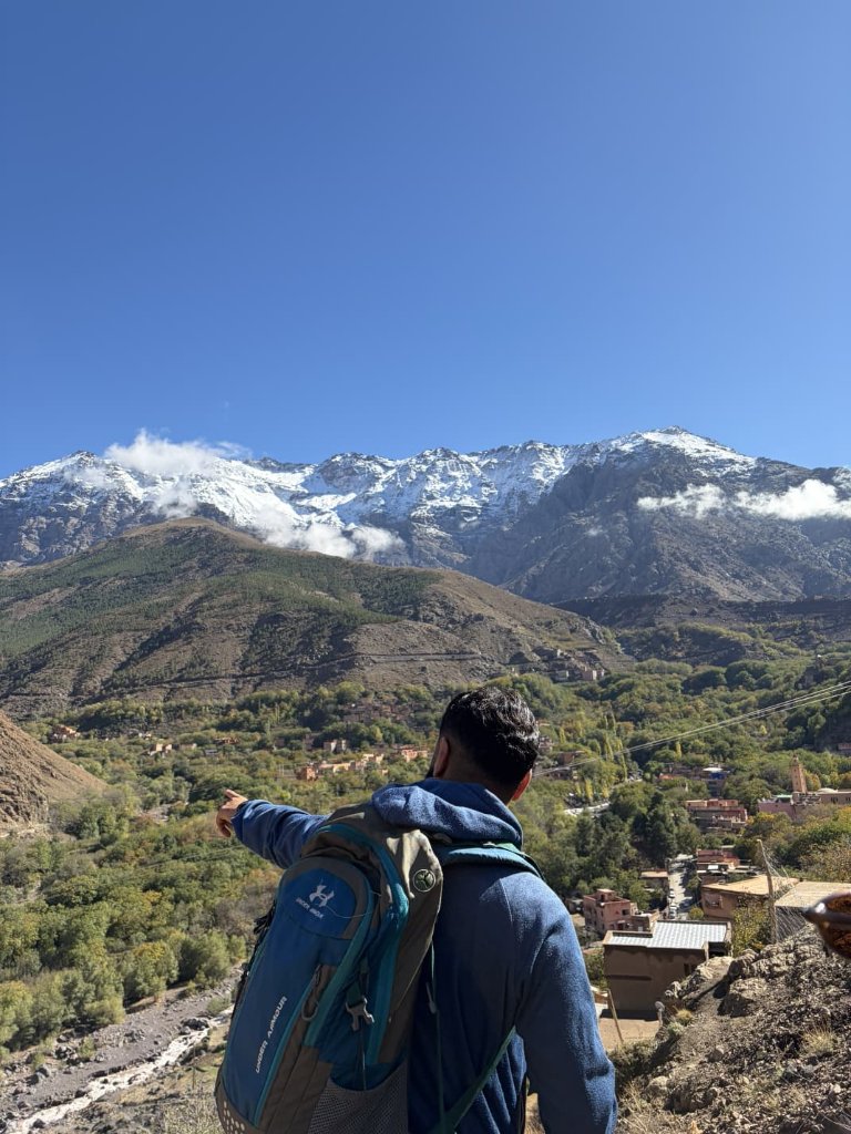 A trekker with a backpack on a rocky High Atlas outcrop, facing snow-capped peaks