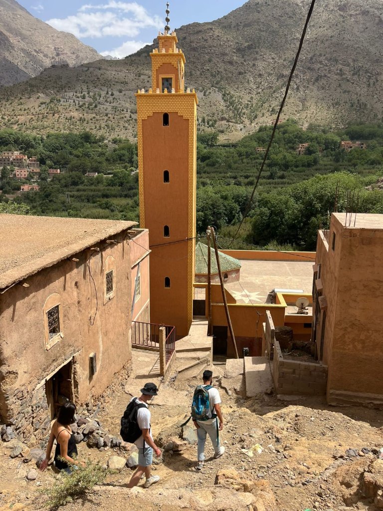Two travellers walking with their guide down a village path toward the mosque in Imlil