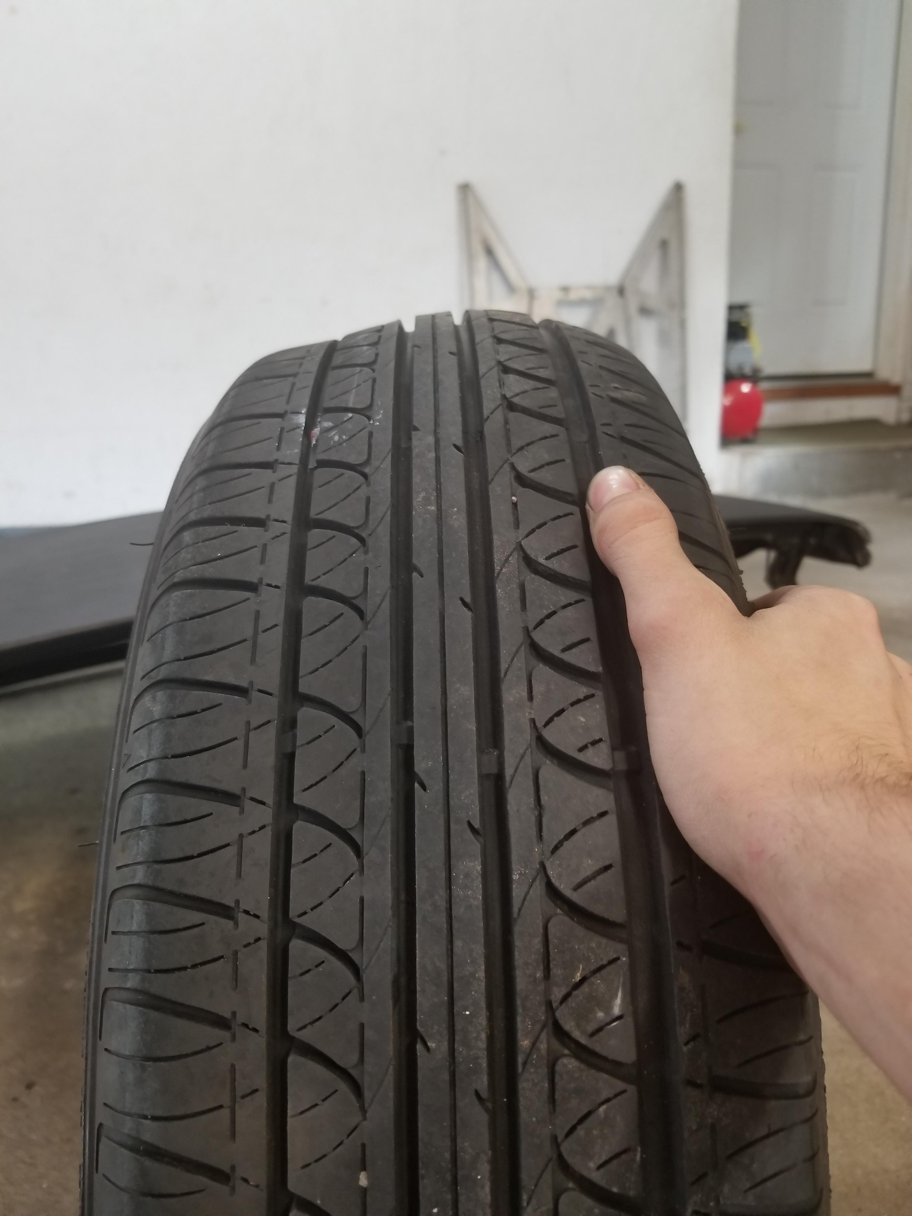 Top-down view of a tyre tread with a thumb pressing near a damaged edge, showing exposed fibrous casing material.