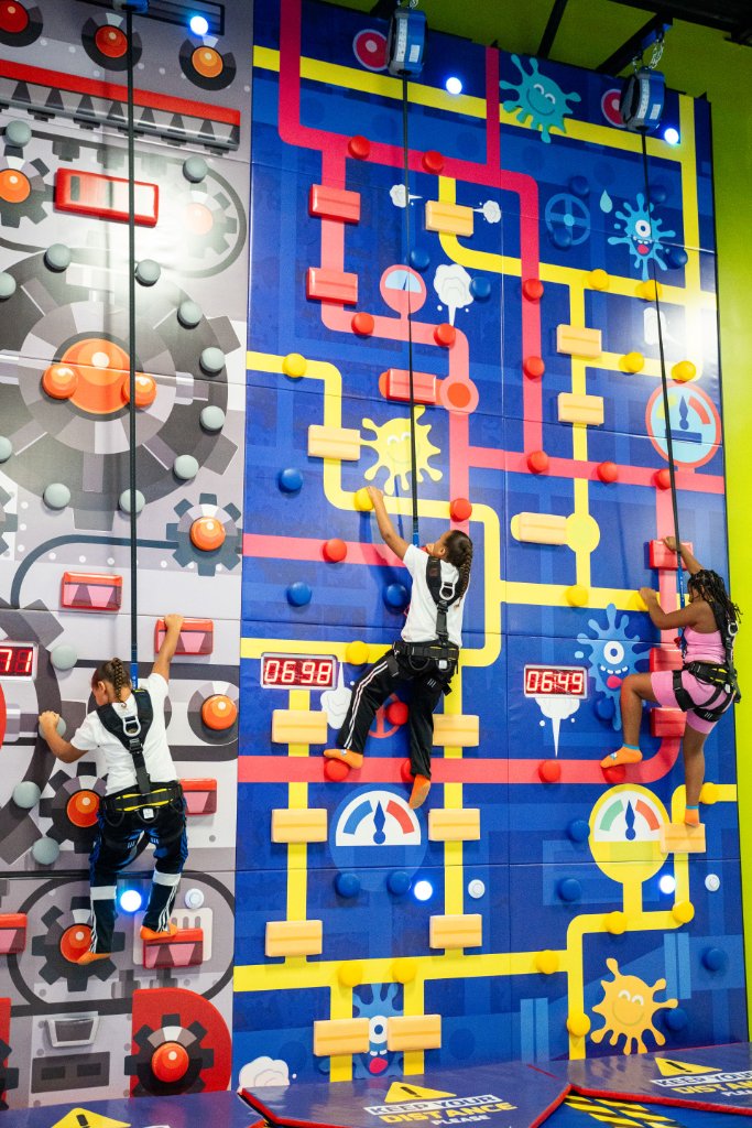 Children in safety harnesses climbing a colorful tech-themed climbing wall with digital timers