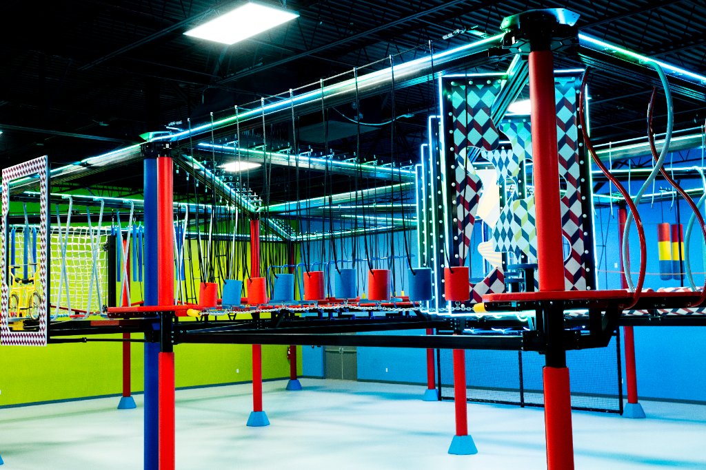 Elevated indoor ropes course with hanging red cylinders, blue blocks, and climbing nets
