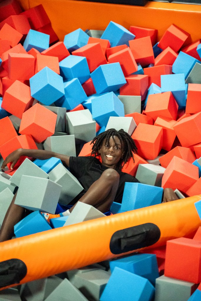 A happy child reclining in a colorful foam pit filled with orange, blue, and grey foam cubes