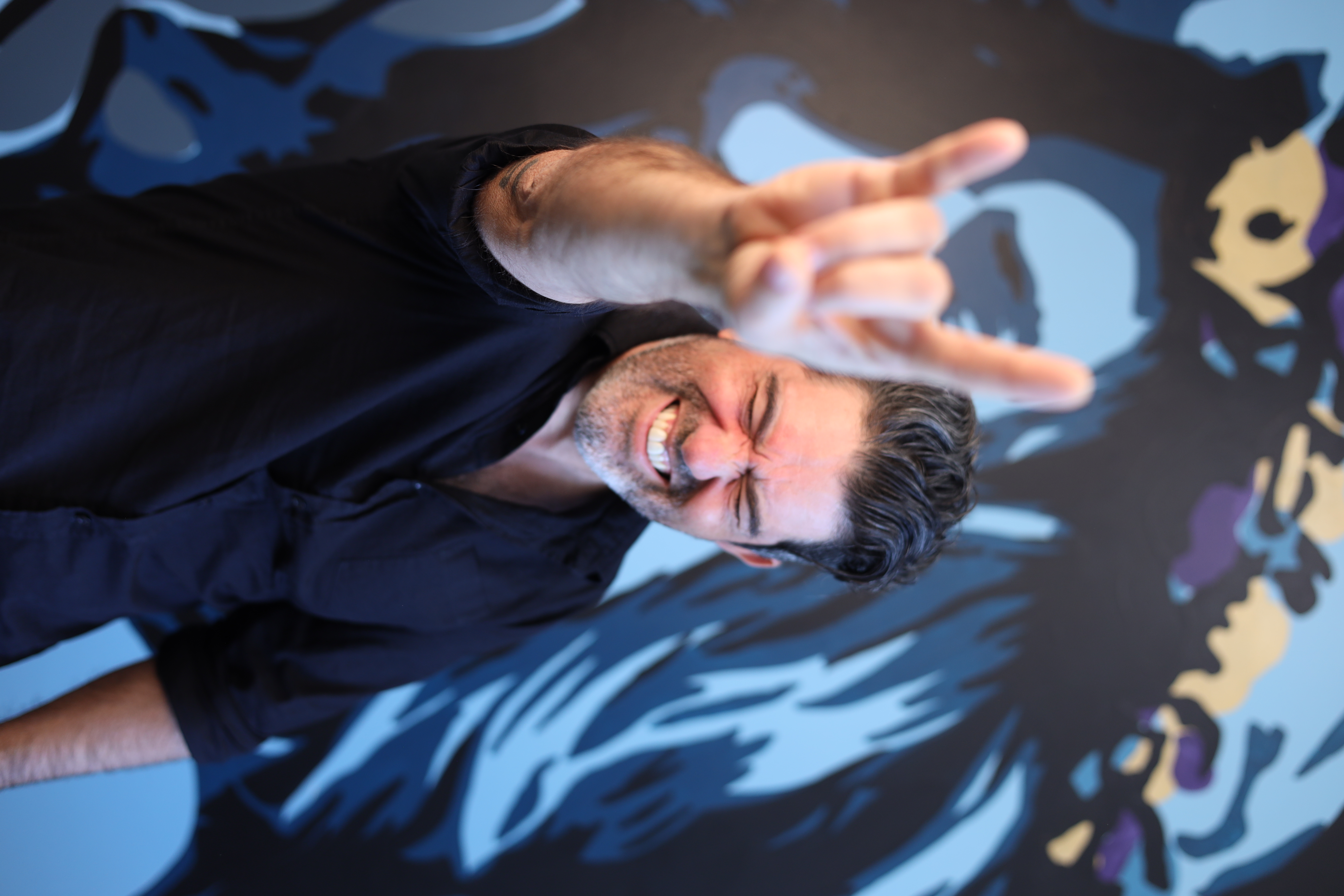 Domenic Fusca smiling broadly with his hand raised in a rock and roll gesture in front of a colourful abstract mural