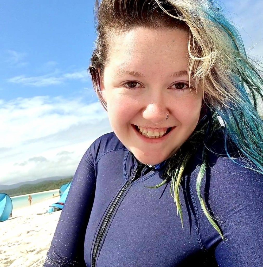 Naomi, founder of The Long Haul Careers, smiling at the beach in a navy wetsuit with blue-teal hair, photographed outdoors in natural light