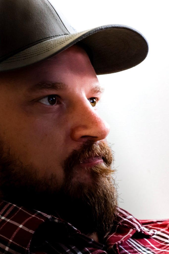 Profile portrait of bearded man in baseball cap and red plaid flannel shirt looking off-camera against white background