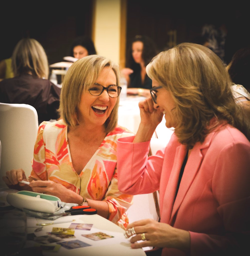 Two women laughing together at a white-clothed event table, one in patterned coral blouse and one in pink blazer