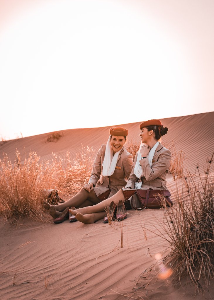 Two flight attendants sitting on a sand dune laughing together at golden hour