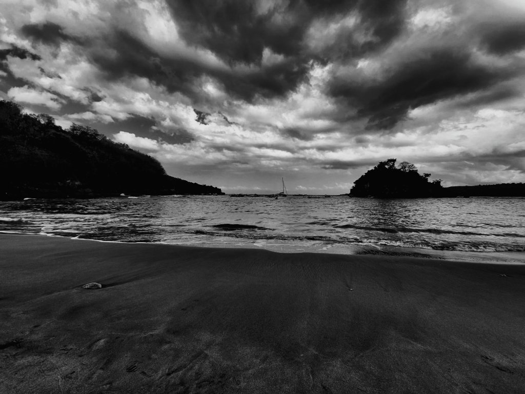 Black and white seascape with dramatic storm clouds, beach, and sailboat on horizon