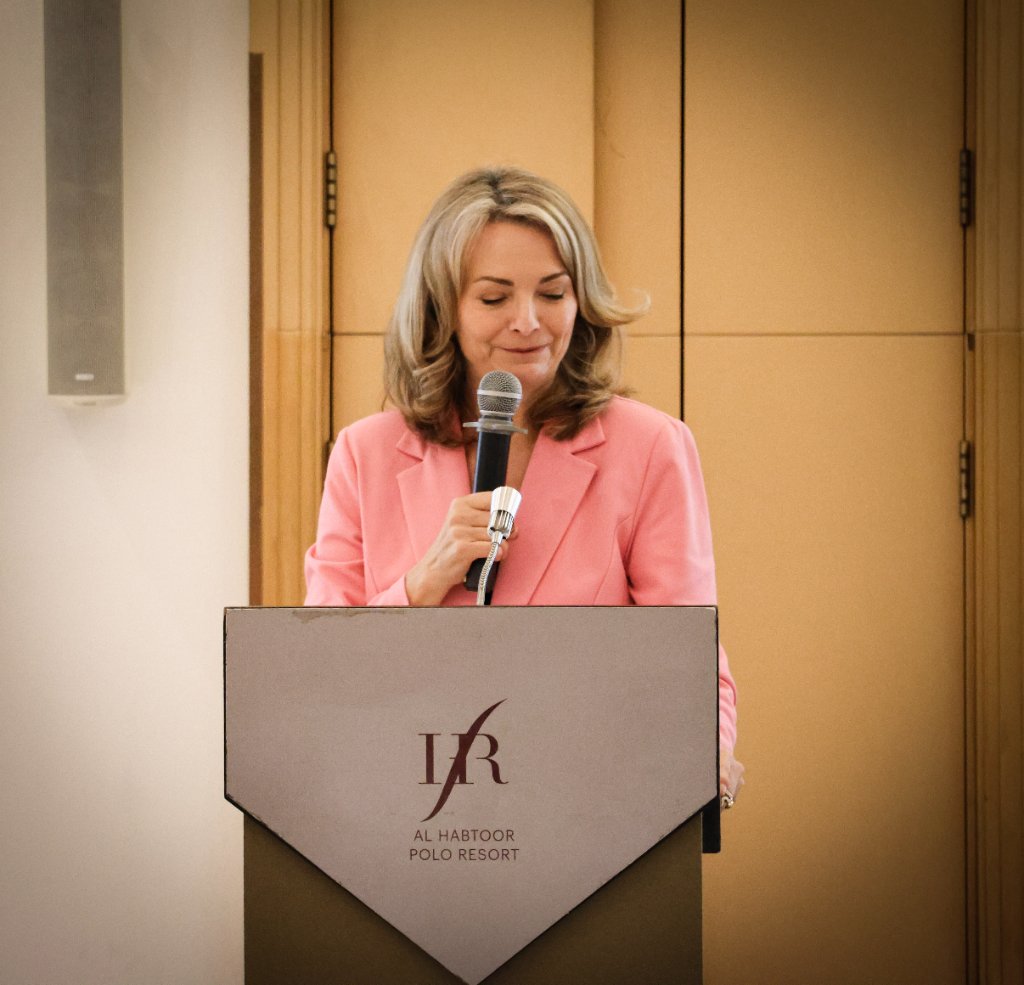 Woman in bright pink blazer speaking at podium with Al Habtoor Polo Resort logo at corporate event