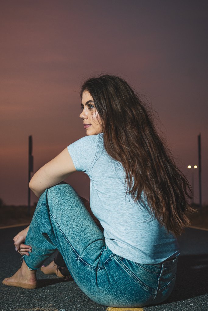 Woman sitting barefoot on an asphalt road at dusk with warm horizon glow