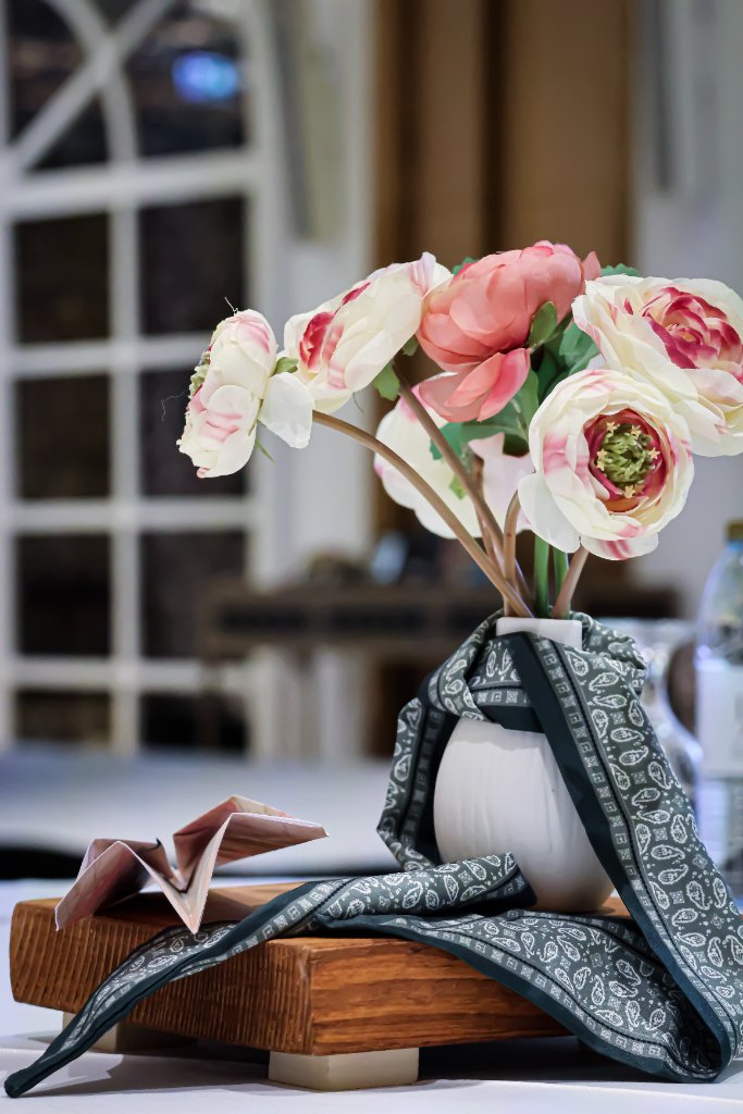 White vase with cream and coral flowers, green patterned scarf wrap, and origami map paper plane on wood riser