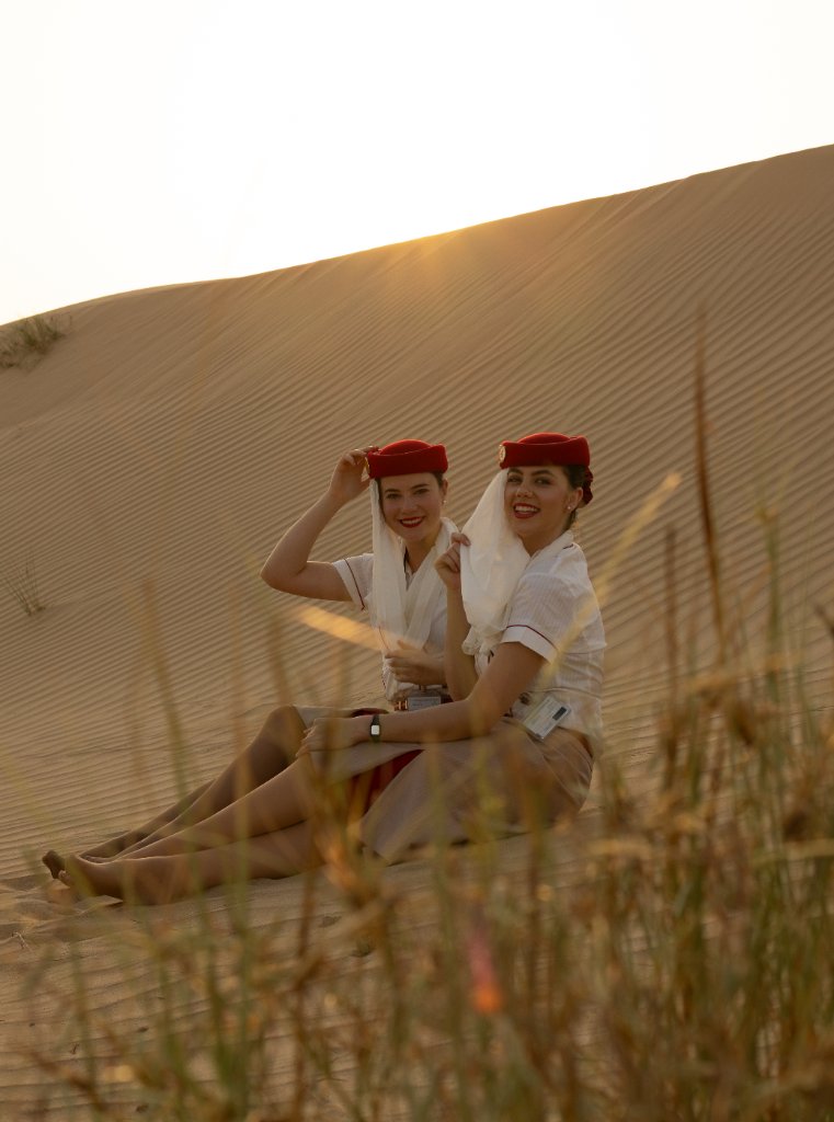 Two flight attendants seated in the desert adjusting their red hats at sunset