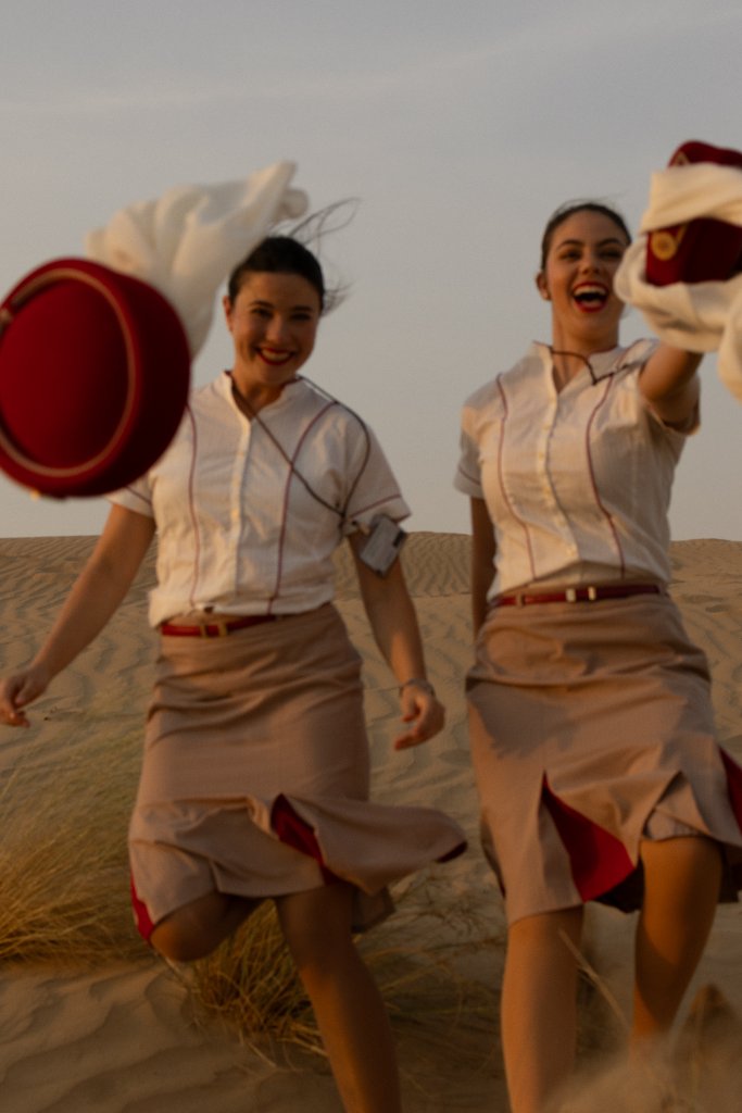 Two flight attendants running across a sand dune holding their red hats at golden hour