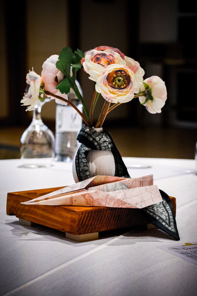 White ceramic vase with blush ranunculus flowers, dark patterned scarf, and folded map paper airplanes on wooden base