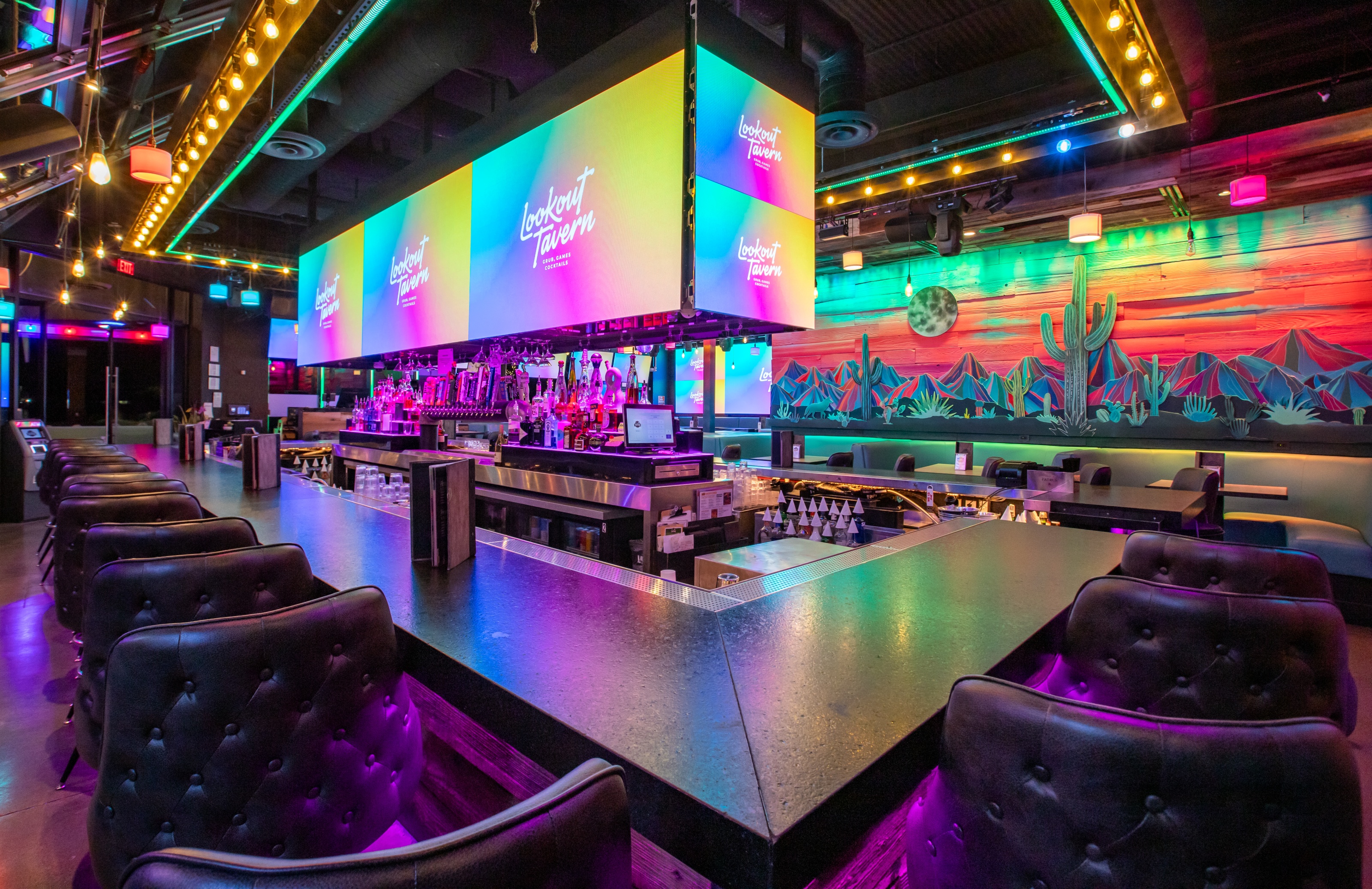 A large U-shaped bar with dark leather chairs and neon-lit desert murals under screens displaying Lookout Tavern logos.