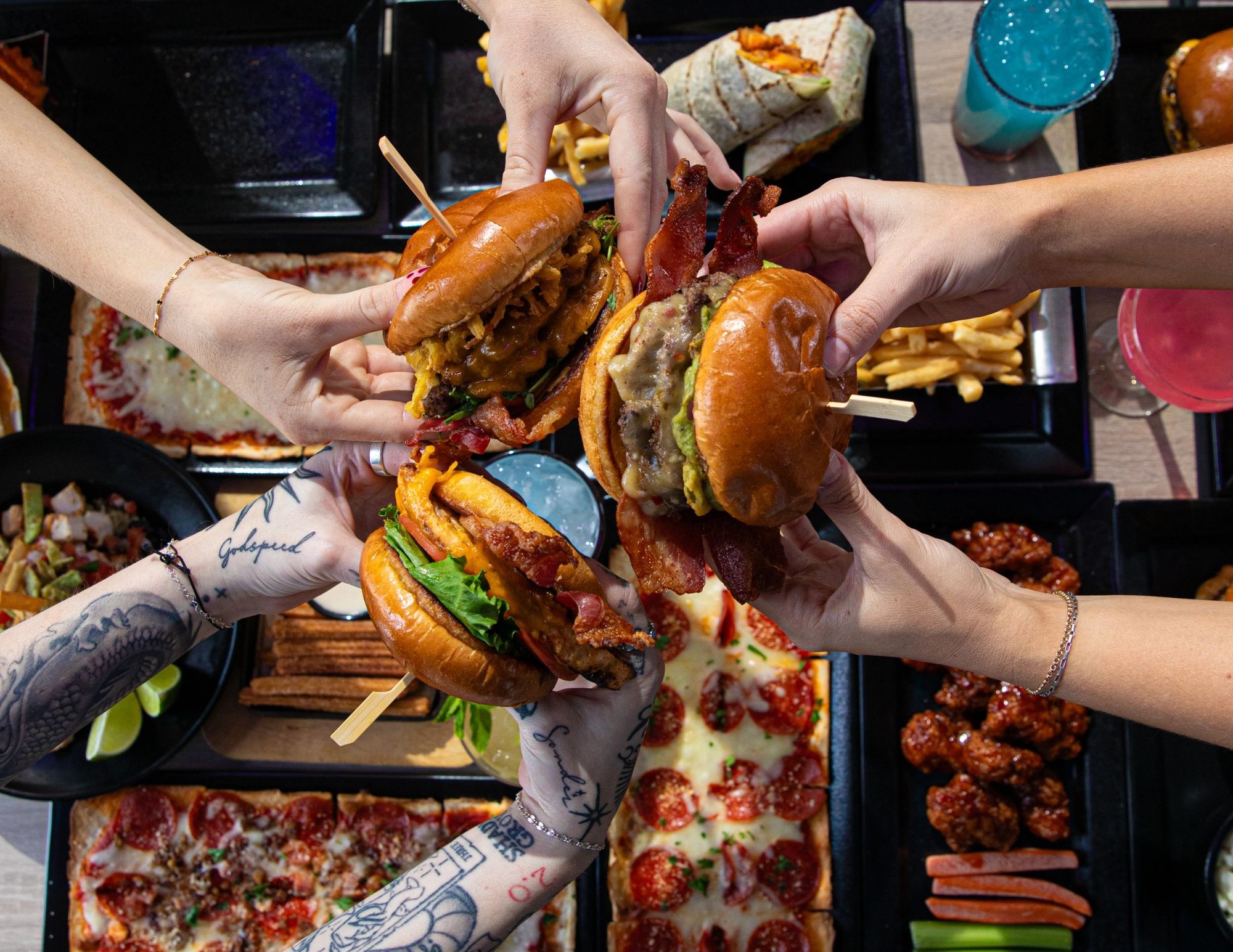 Hands grabbing smash burgers over pizza, wings and wraps