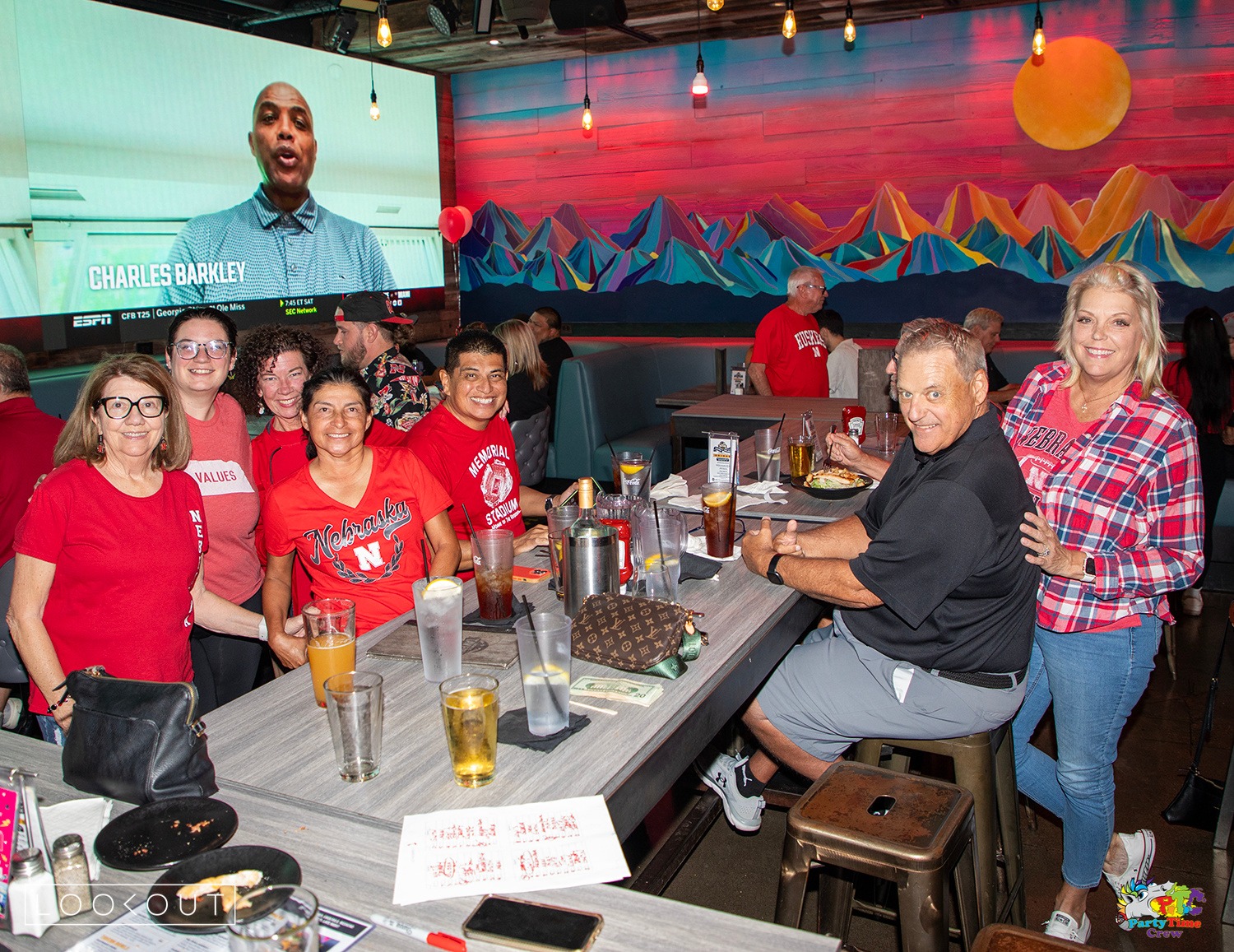 Huskers fans in red at a long table under the desert mountain mural, college football on the LED wall