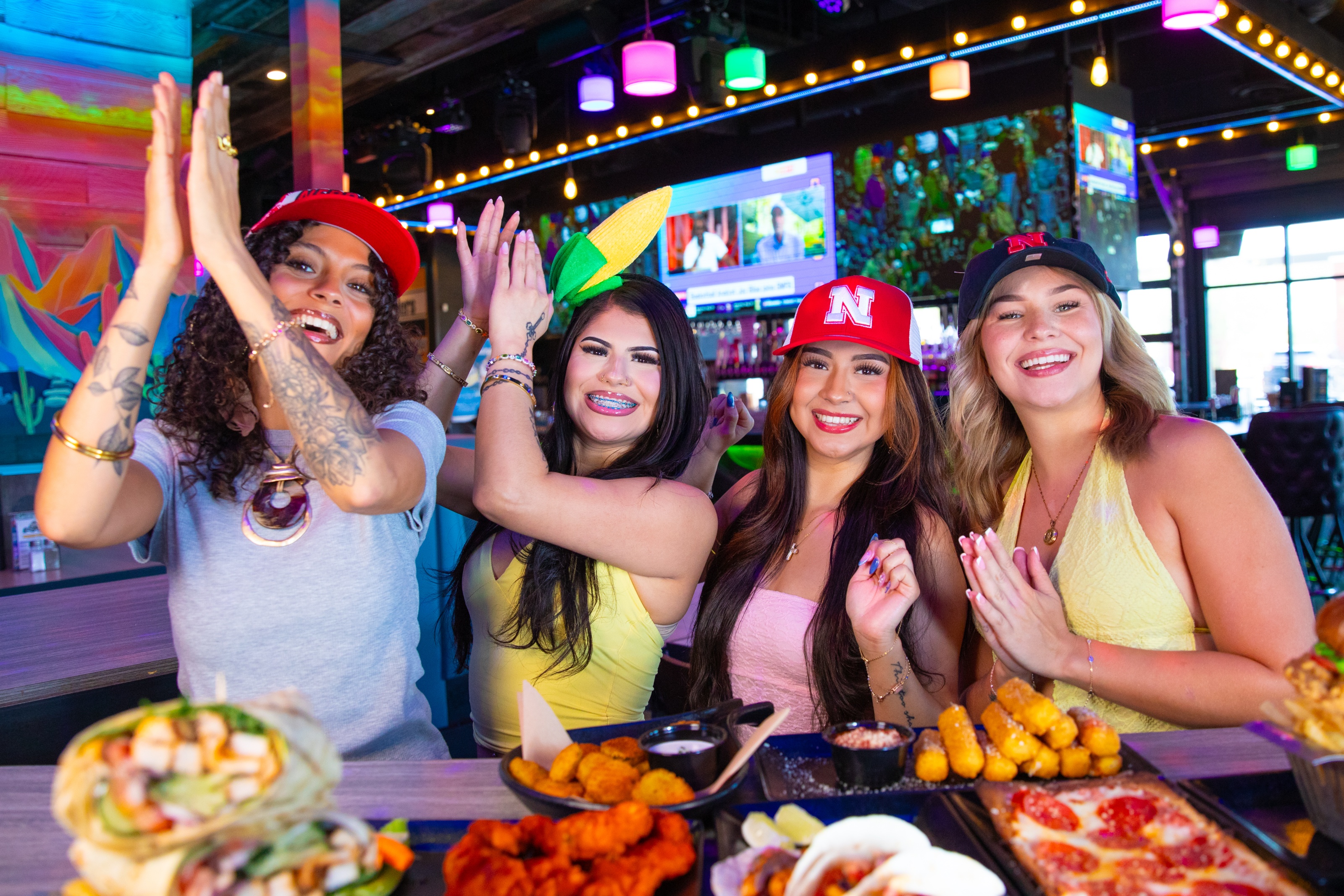 Four young women smiling and clapping while sitting at a table filled with food at a sports bar with large TVs.