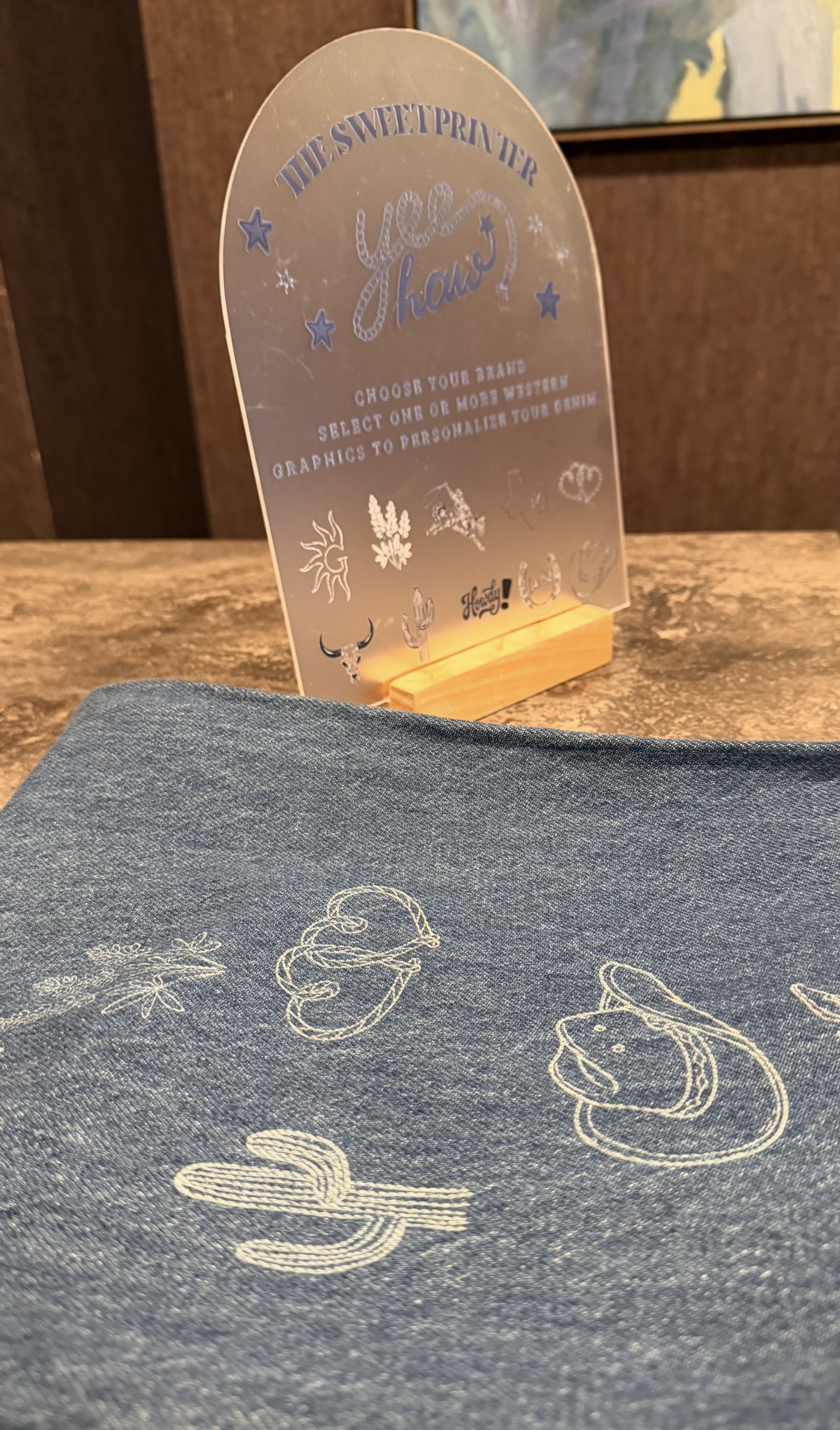 A frosted acrylic 'Choose Your Brand' sign listing western graphics for live denim engraving, with personalized embroidered denim beside it