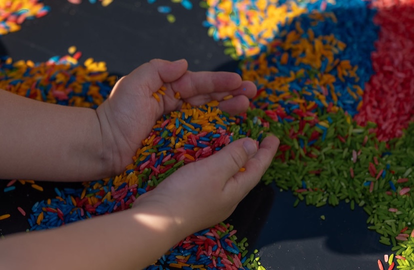 Hands cupping colourful dyed rice in a sensory tray
