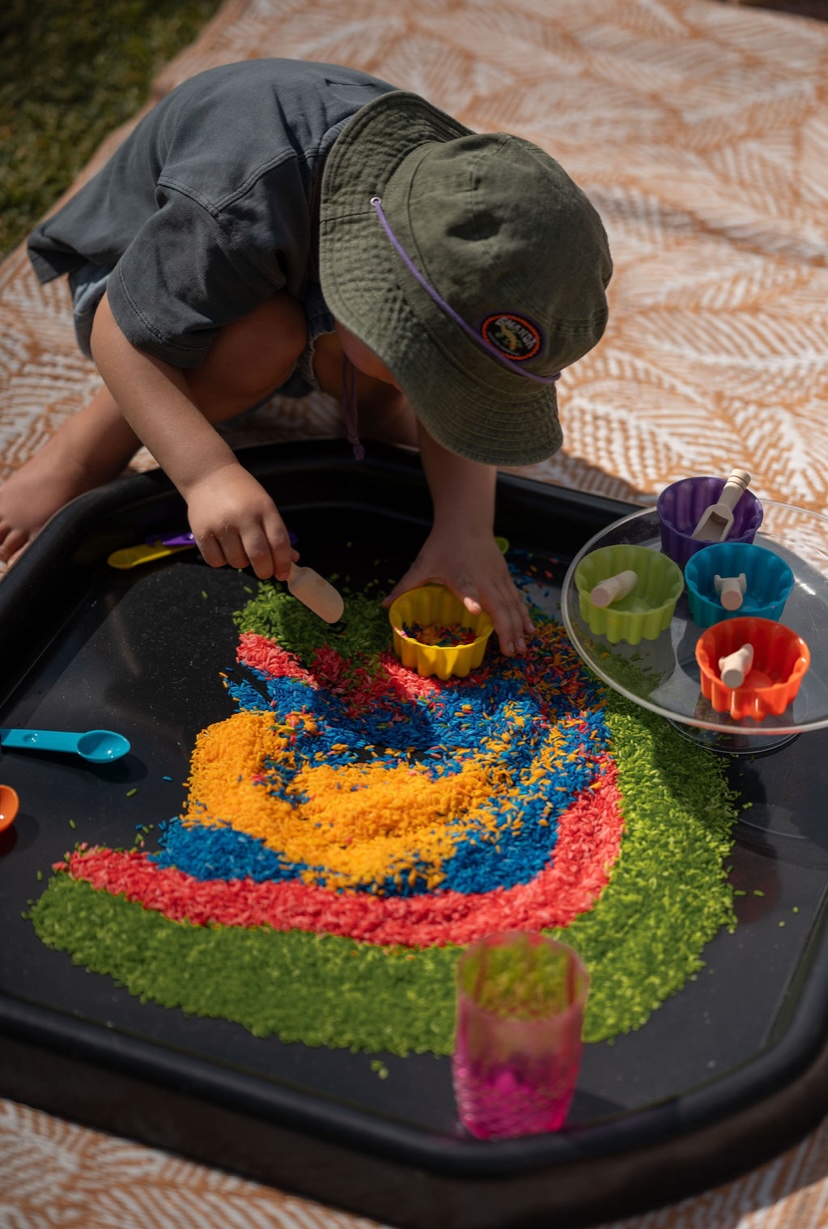 Child scooping rainbow rice on a sensory tuff tray