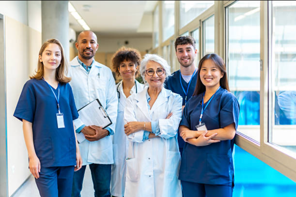 A diverse team of six doctors and nurses in blue scrubs and white lab coats posing together and smiling in a bright hospital corridor