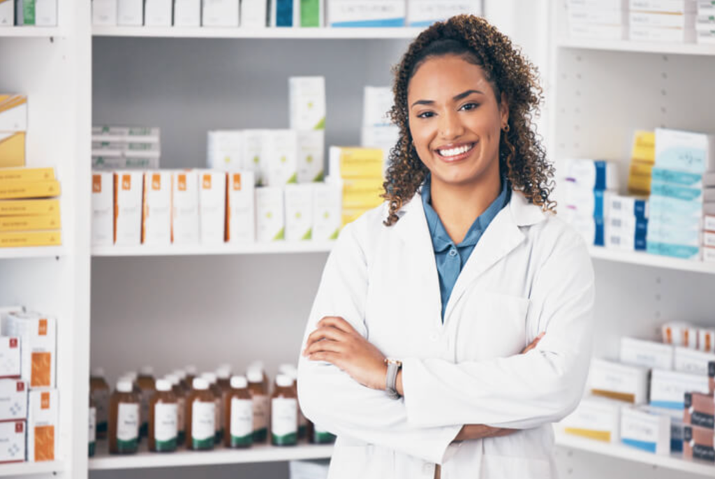 Smiling pharmacist with curly dark hair in a white lab coat and blue shirt, standing with arms crossed in front of shelves stocked with pharmacy products and medicine bottles