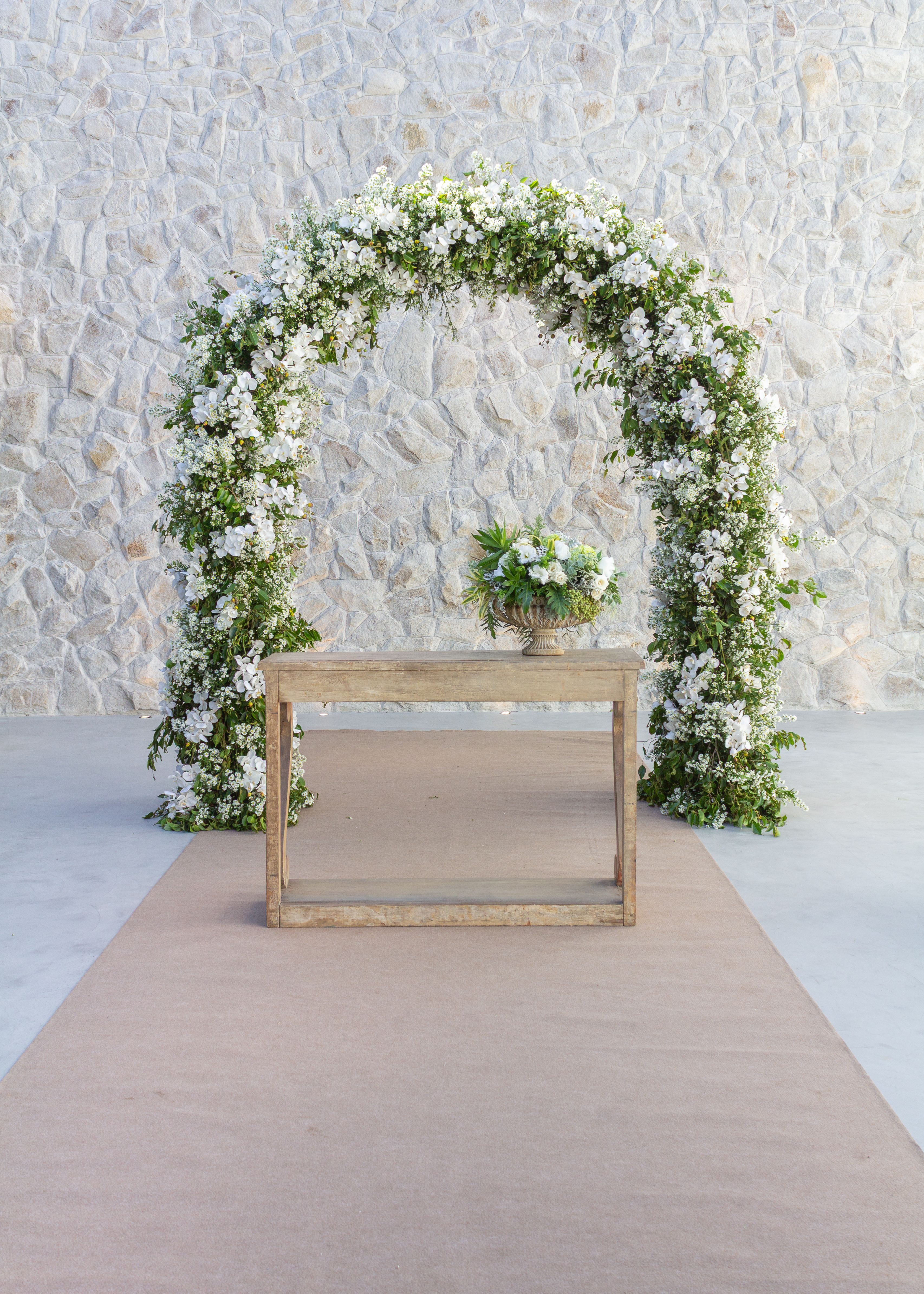 Wedding ceremony arch of white blossoms and lush greenery against a stone wall - Kalea Arrangements