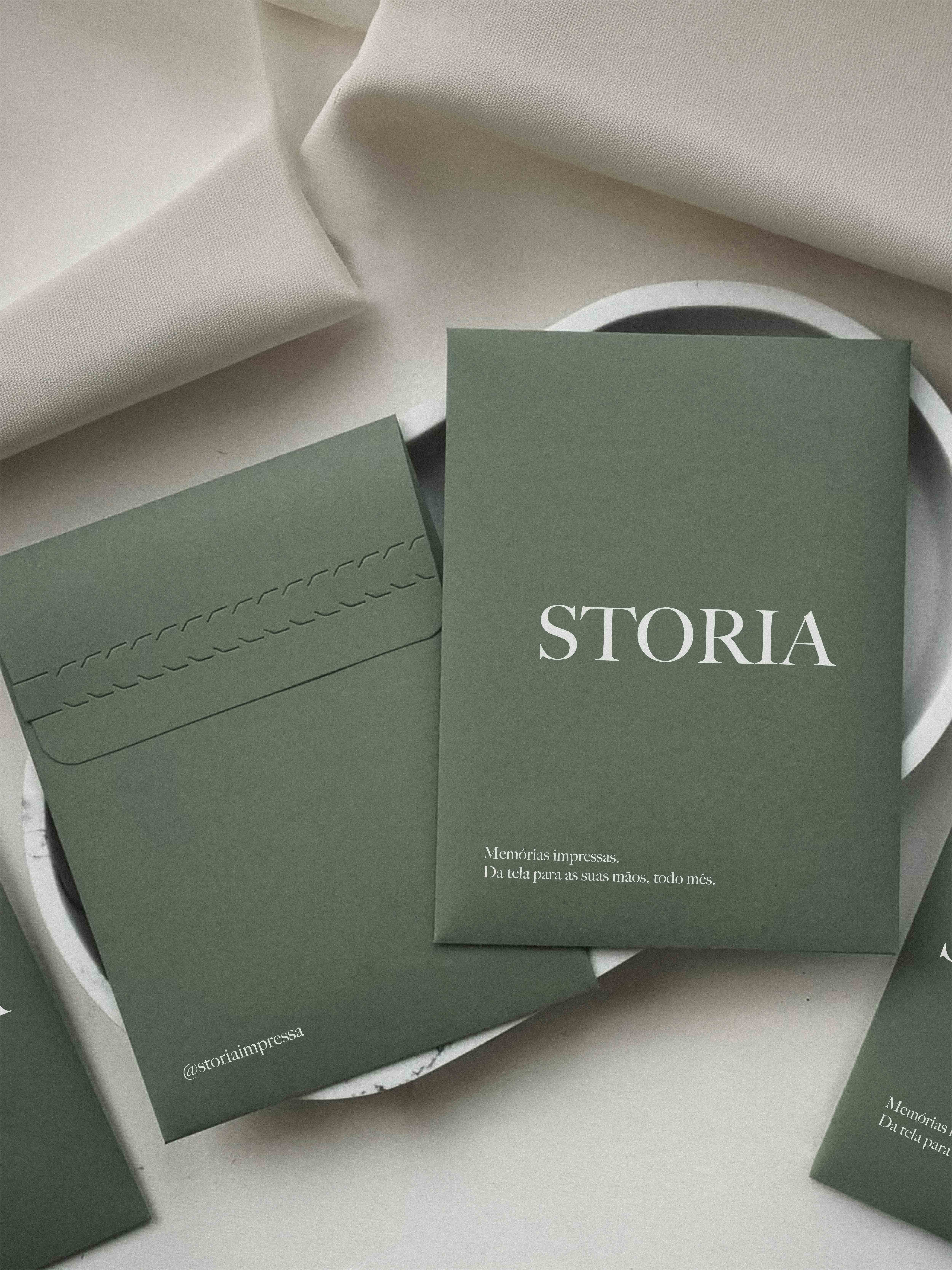 Envelope verde sage da Storia com o nome da marca e a frase Memórias impressas, sobre superfície de mármore