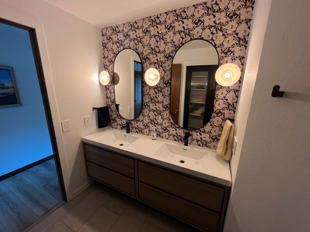 Double vanity straight-on — floral wallpaper, globe sconces, dual oval mirrors, matte black faucets