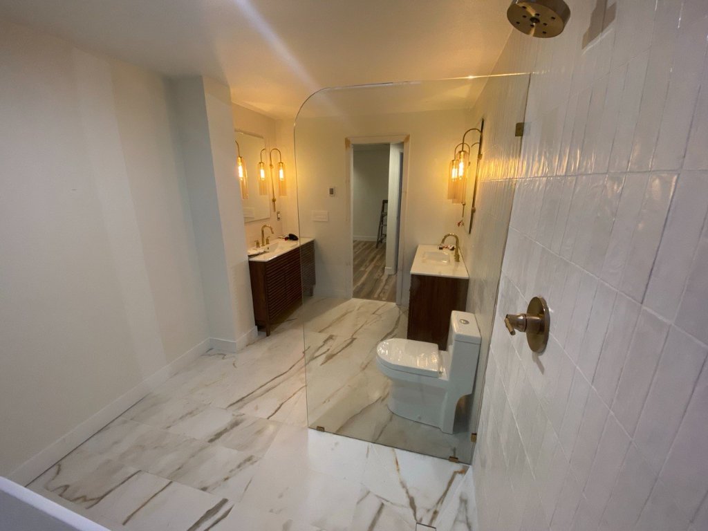 Full suite view from inside curbless shower — dual walnut vanities, brass sconces, Calacatta marble floor