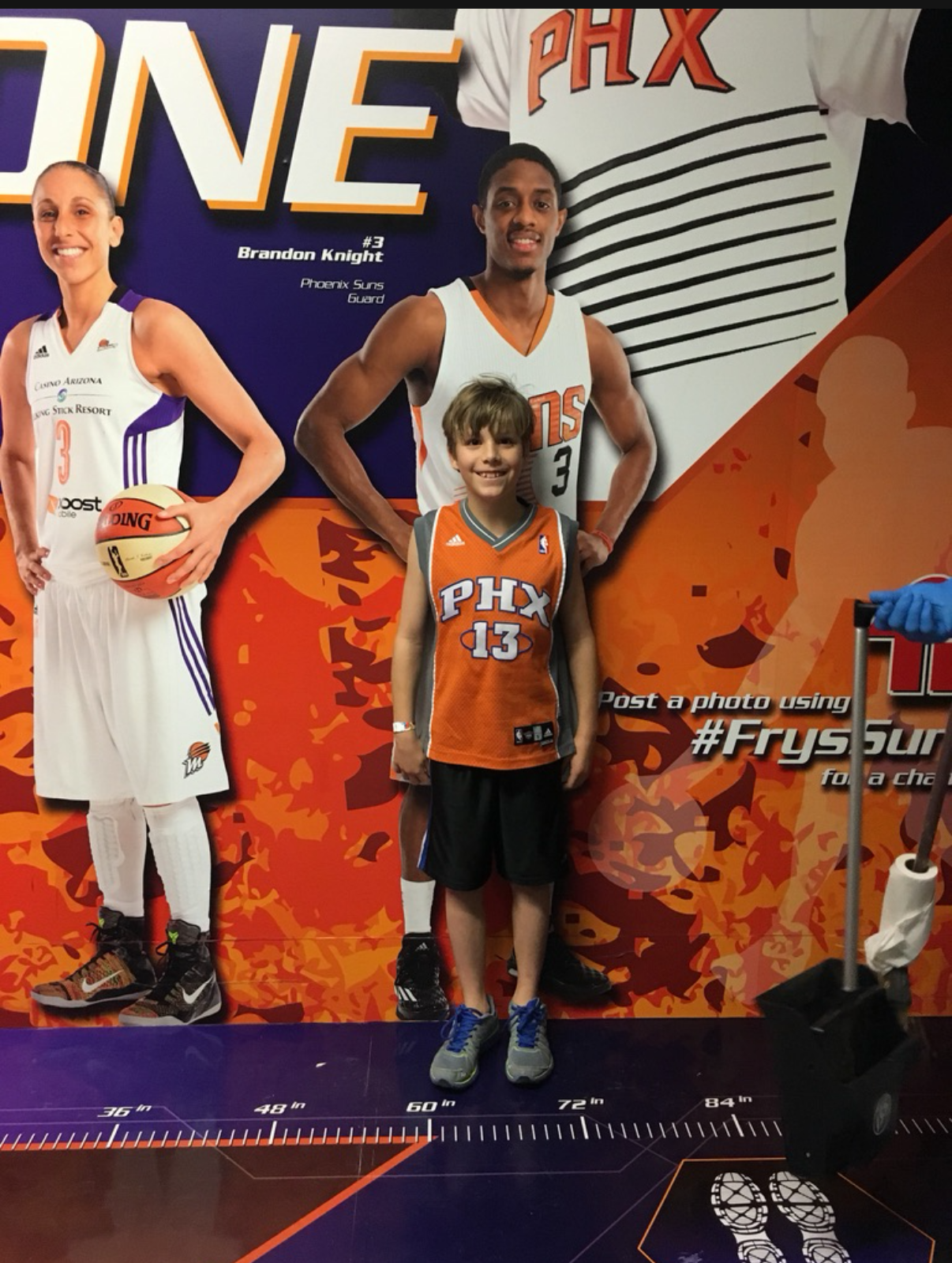 A young Miles Wallace in a Phoenix Suns jersey standing in front of a Brandon Knight promotional backdrop at a Suns event.