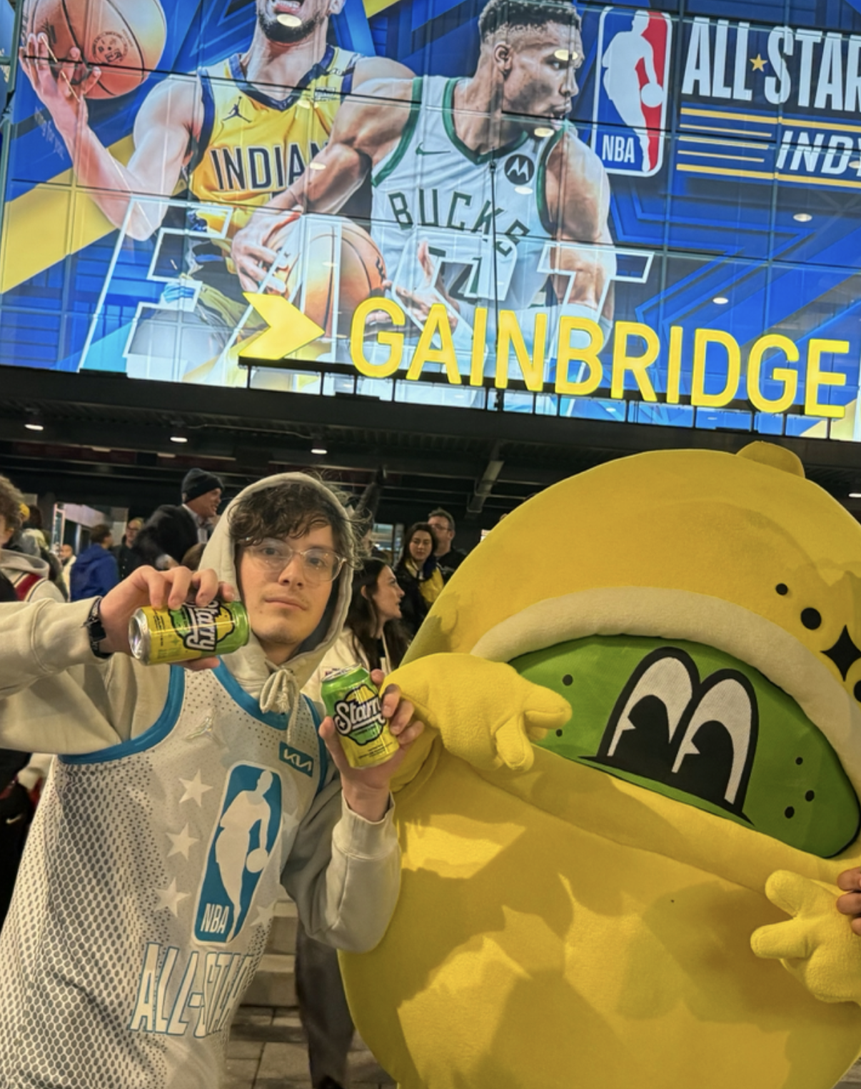 Miles Wallace holding cans of Starry soda with a mascot at an NBA All-Star event, with arena signage visible behind them.