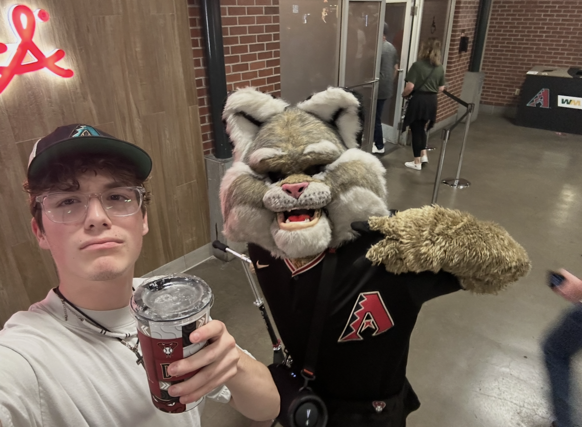 Miles Wallace taking a selfie with a mascot in a stadium concourse, holding a beverage cup.