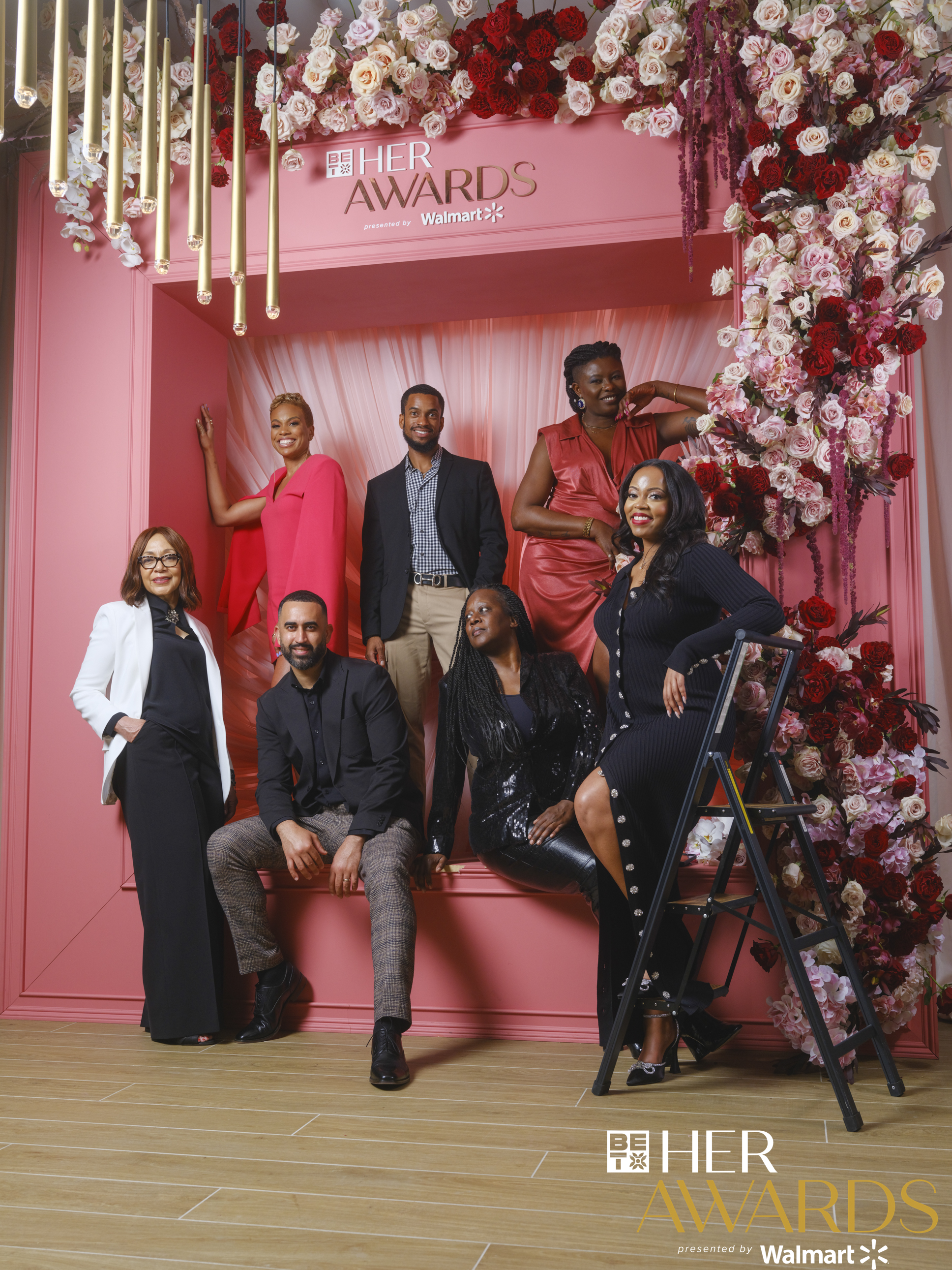 Group of seven guests posed before a pink floral backdrop with gold pendant lighting at the BET Her Awards