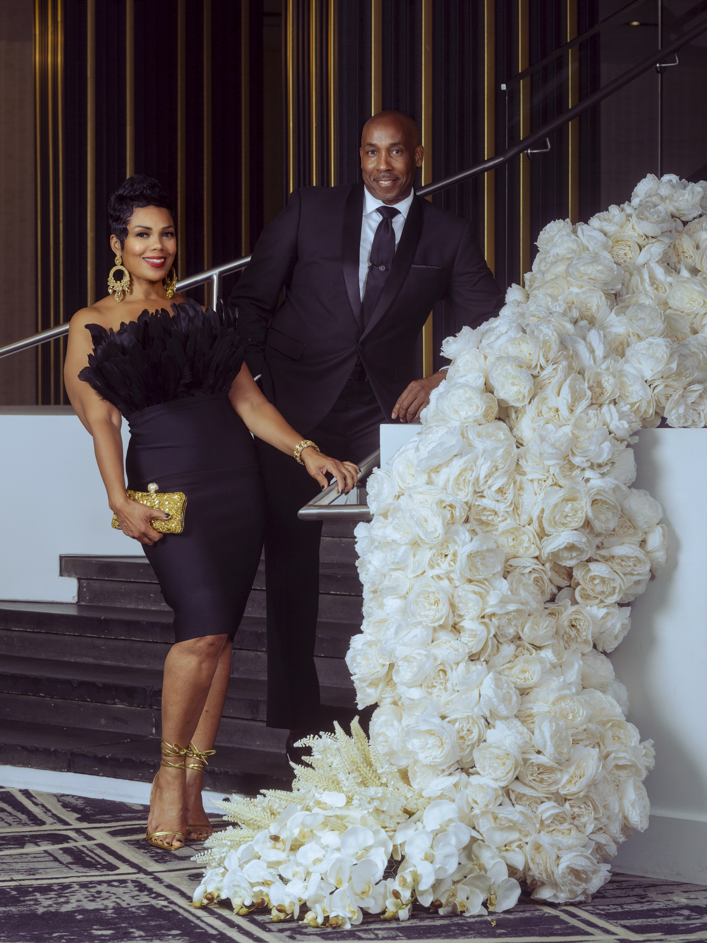 Couple in formal attire posed beside a cascading white floral installation on a staircase