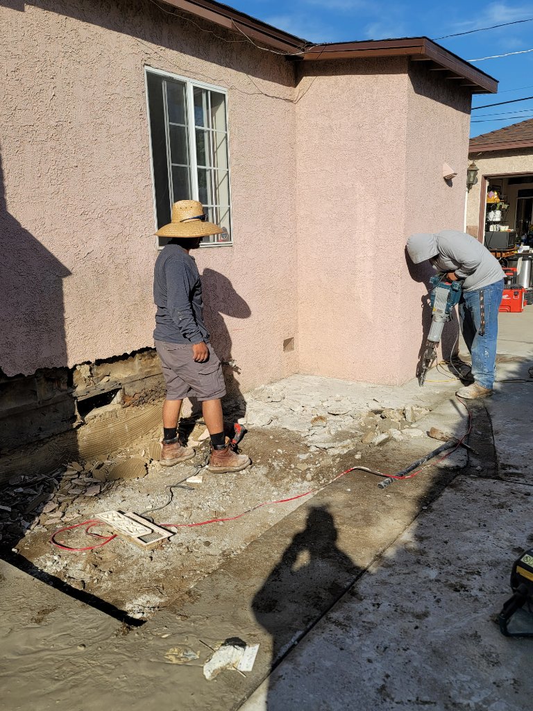 Worker kneeling jackhammering concrete next to pink stucco wall — demolition for room addition California