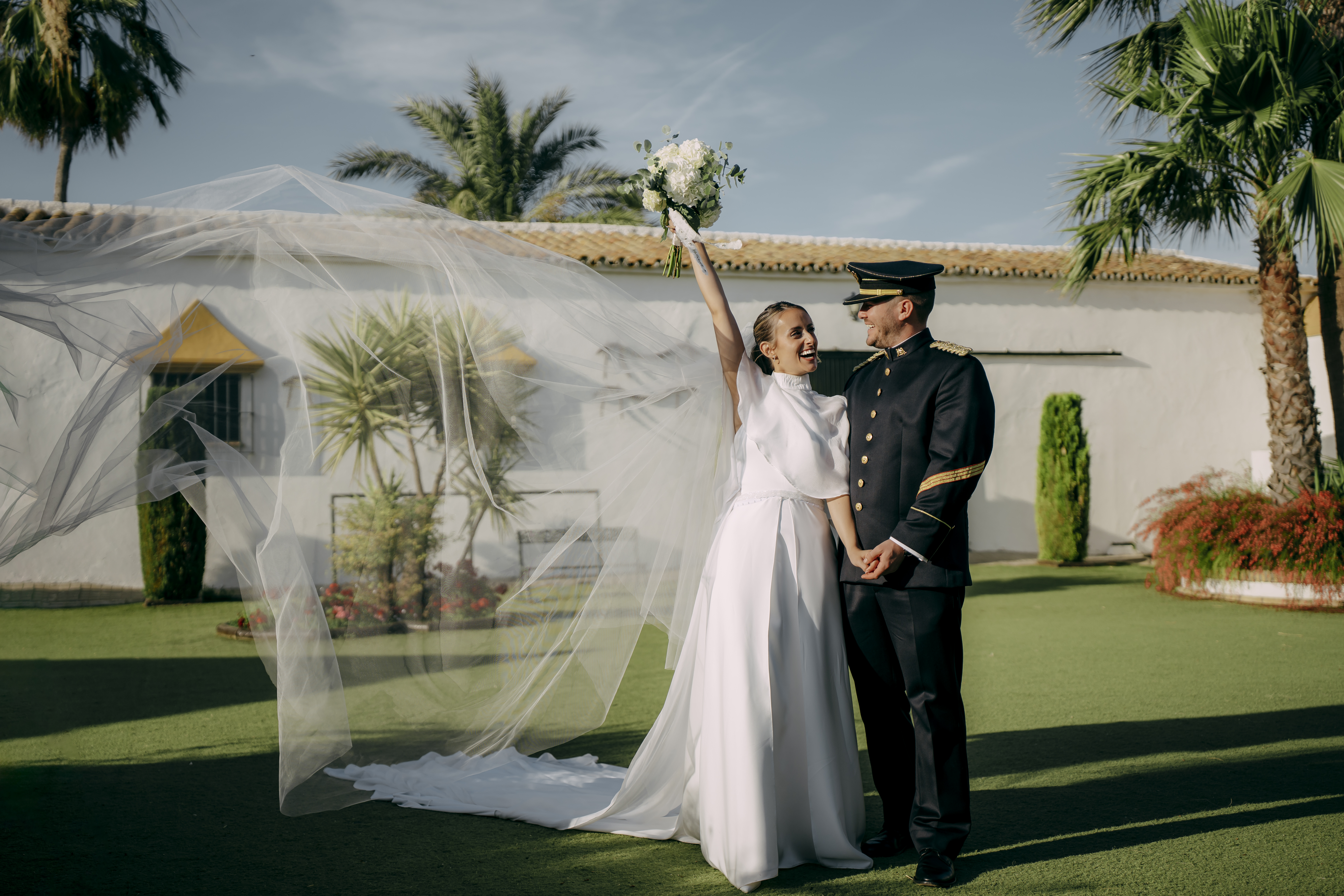 Novia levantando el brazo con el velo al viento junto al novio