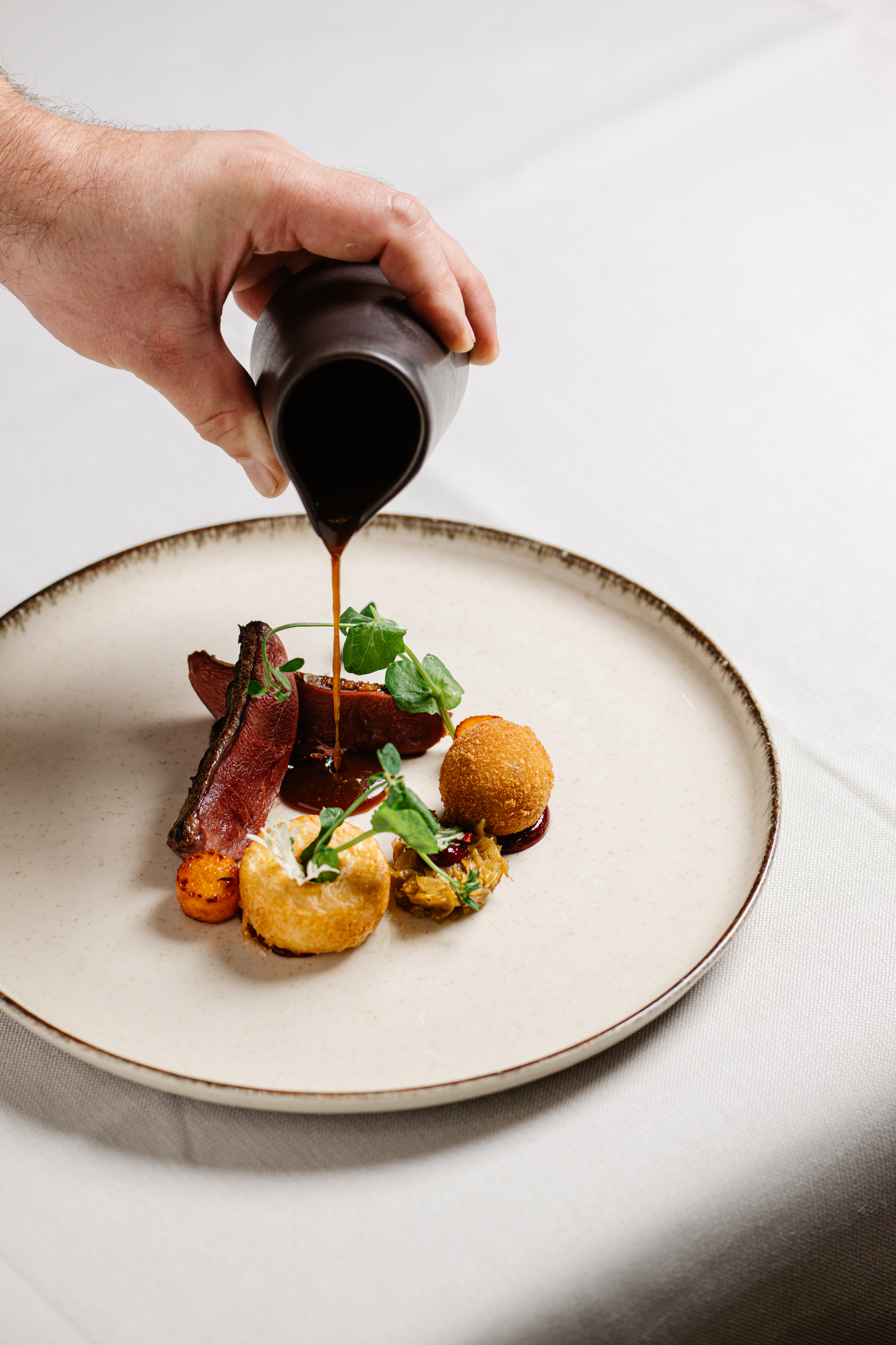 Kok die een donkere jus opschenkt over gegaarde Duif en bitterbal van Duif bij Restaurant LUQS