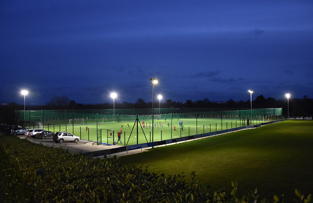 Floodlit football pitch at night with a coach briefing players during a training session