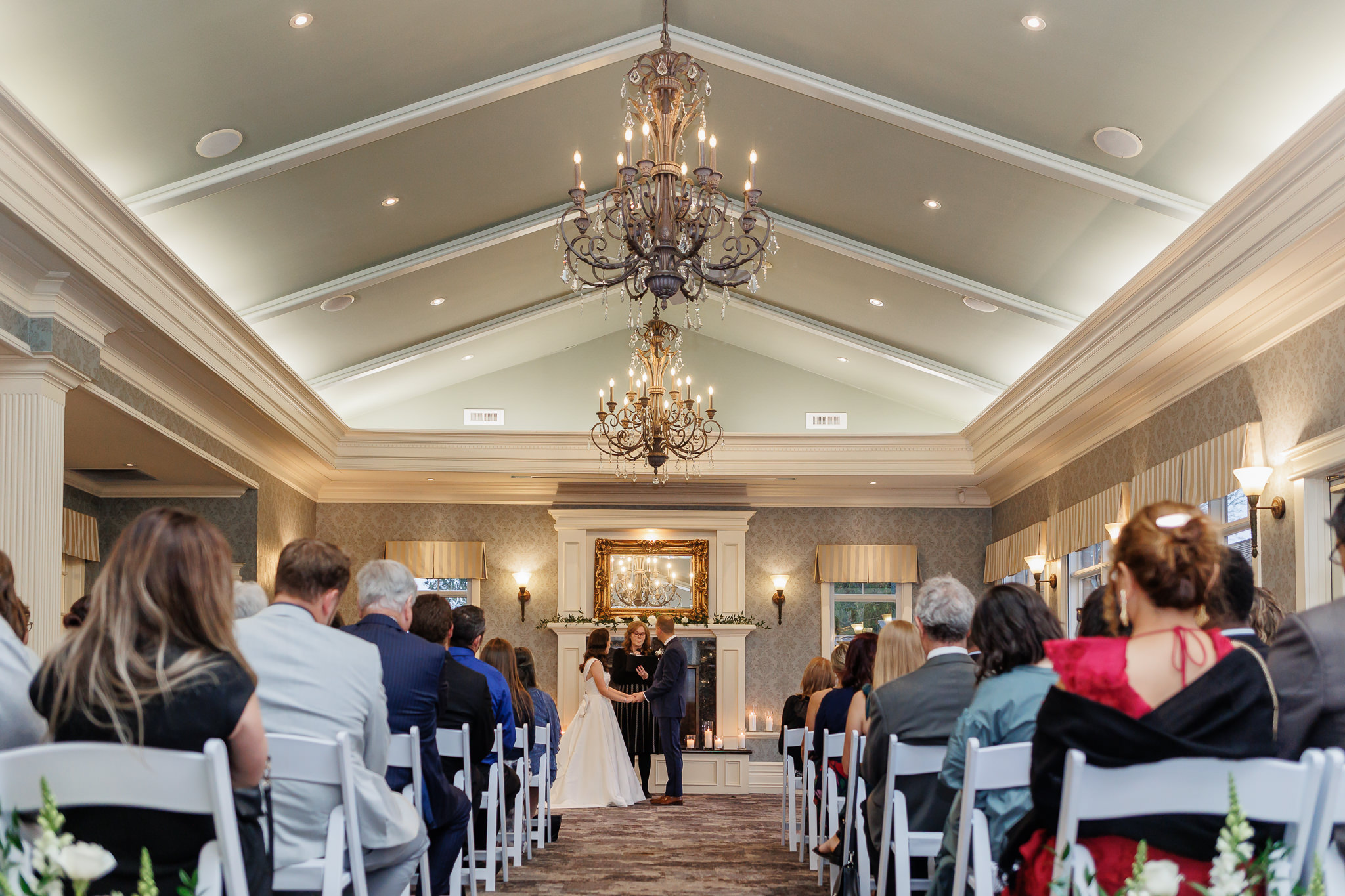 Maureen officiating an indoor wedding ceremony beneath crystal chandeliers with seated guests