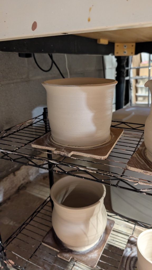 Tall unfired clay cylinder drying on rack