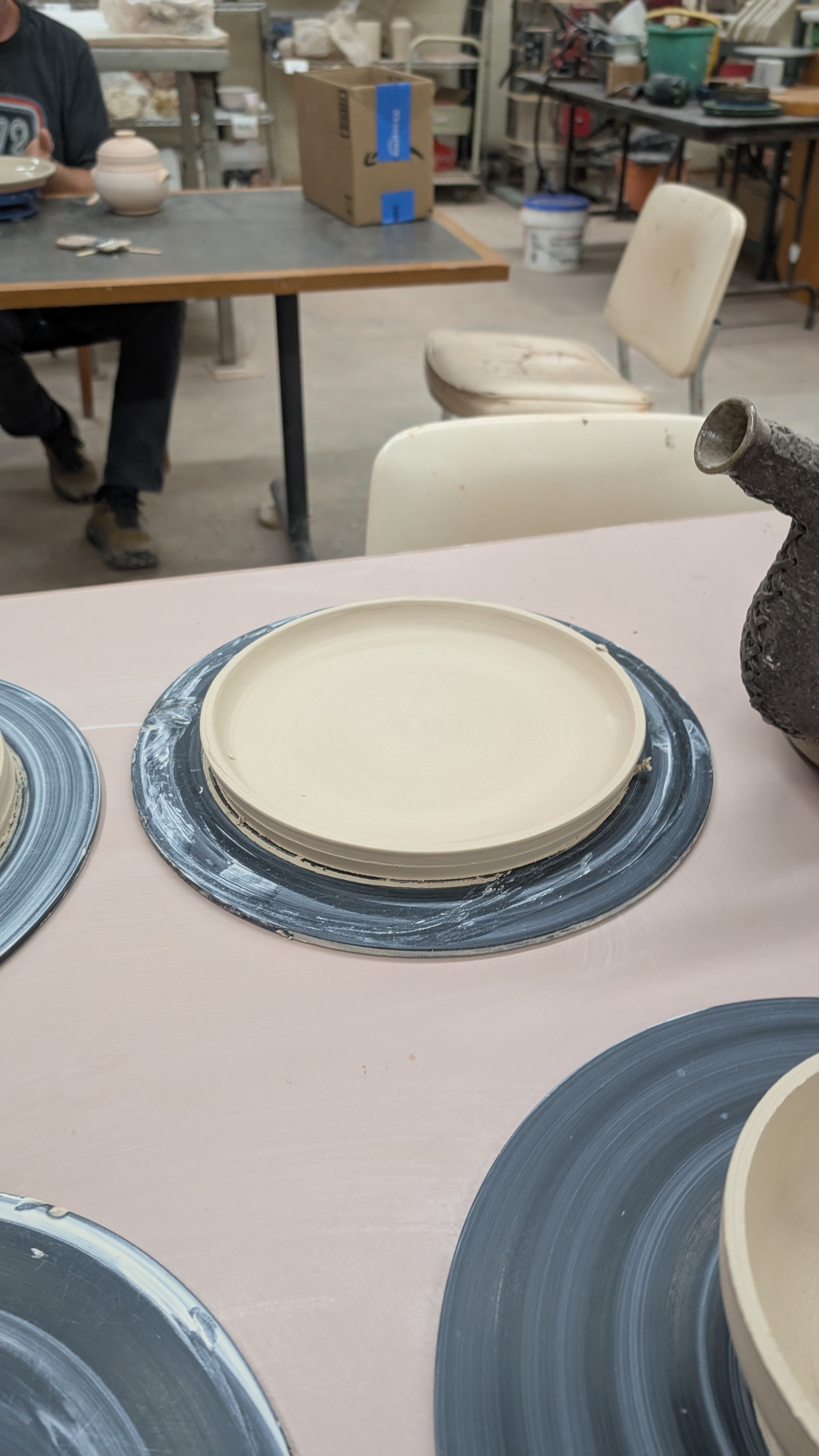 Leather-hard clay plate on throwing bat with teapot nearby