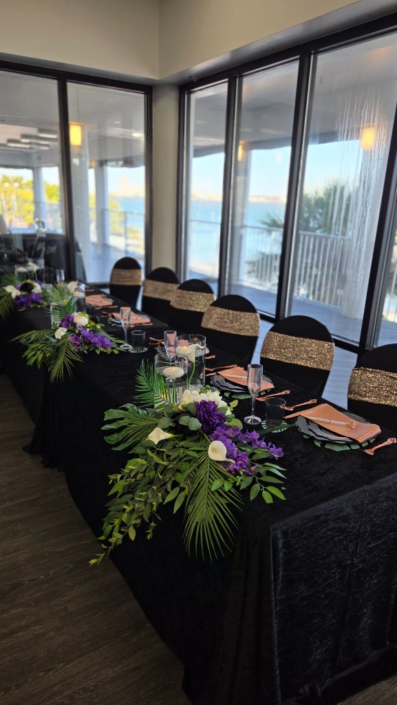 Black velvet banquet table with tropical greenery runner, purple and white florals overlooking the water
