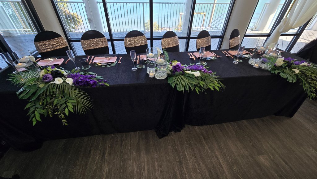 Black velvet head table with tropical purple and white floral garland, gold sequin chair sashes and waterfront view