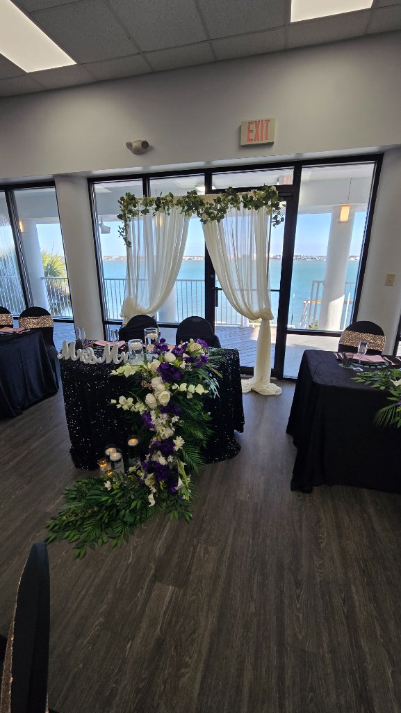 Waterfront wedding venue with white drape arch, black sequin Mr & Mrs sweetheart table and tropical greenery