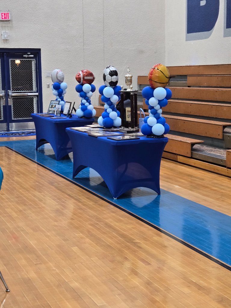 Sports themed balloon towers with baseball football soccer and basketball foil balloons on blue tables in a gym