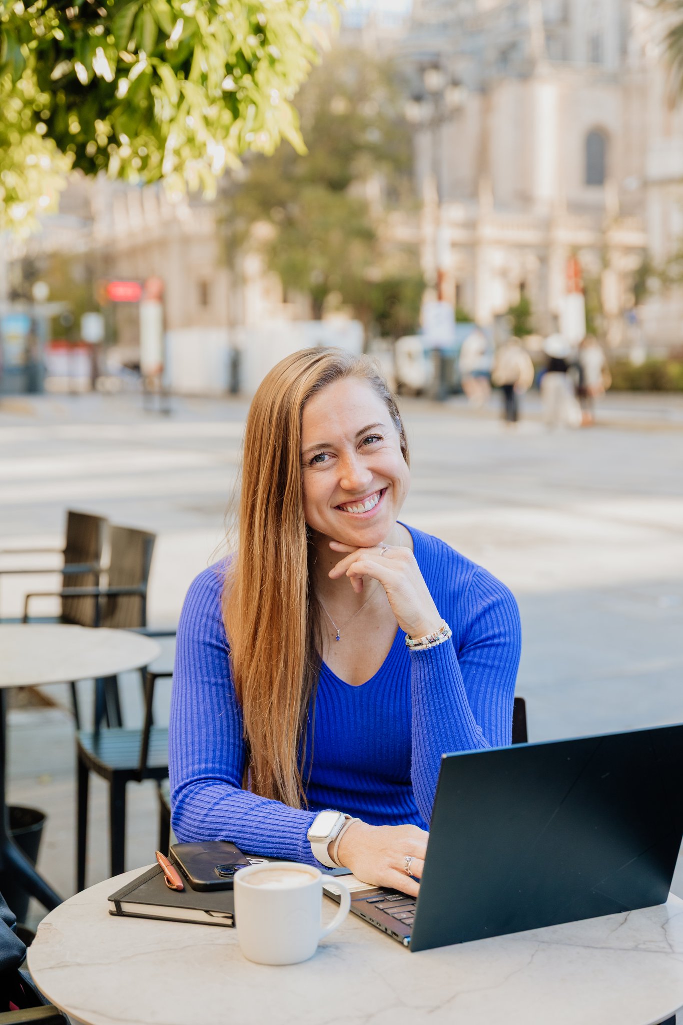 Stephanie Jahn seated at an outdoor cafe table with a laptop in a city plaza, smiling at the camera