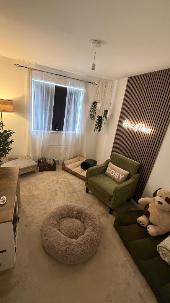 The Retreat Room at House of Paws Retreat — a whole-room view with a soft round dog bed, a moss-green armchair with the COSY cushion, the warm House of Paws neon welcome sign, sheer curtains, hanging plants and a quietly comfortable home-from-home atmosphere