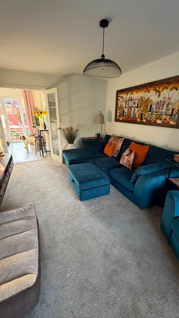 The comfortable living room at House of Paws Retreat — a deep teal L-shaped sofa and ottoman with warm botanical accents, opening onto secure outdoor play through the conservatory
